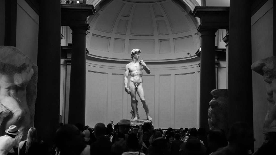 A black and white photo of the Statue of David standing in a gallery under a domed ceiling, with a crowd of people observing it.