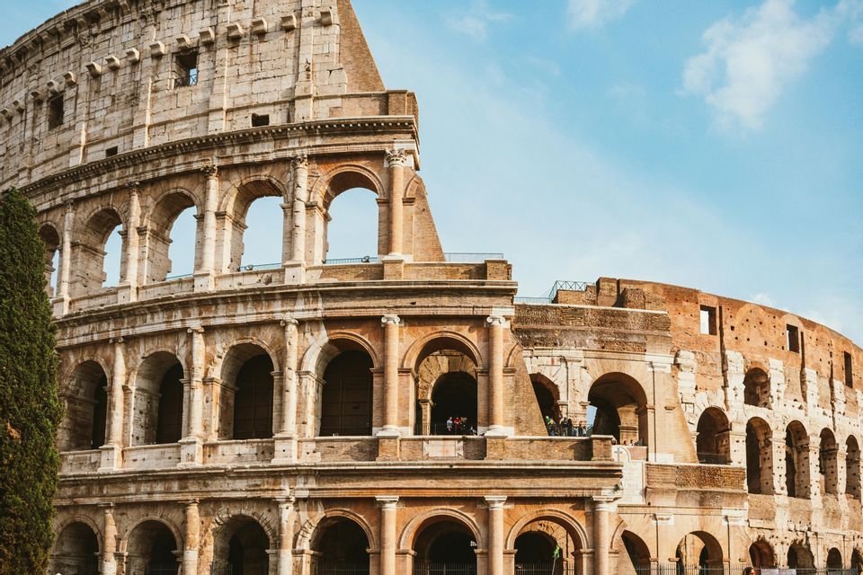 The exterior of a large, ancient stone amphitheater with rows of arches is seen from a low angle against a blue sky.