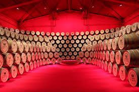 Dans une cave illuminée par une lumière rouge intense, des dizaines de tonneaux en bois sont empilés le long d'une grande paroi incurvée.