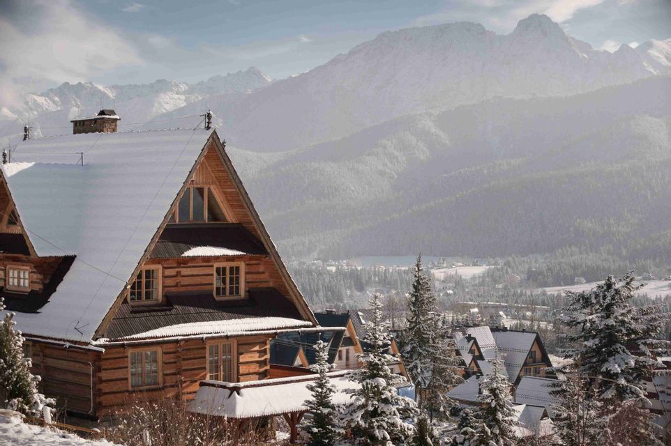 Un chalet de madera con tejado nevado domina un pueblo de montaña y pinos, con grandes picos nevados de fondo.