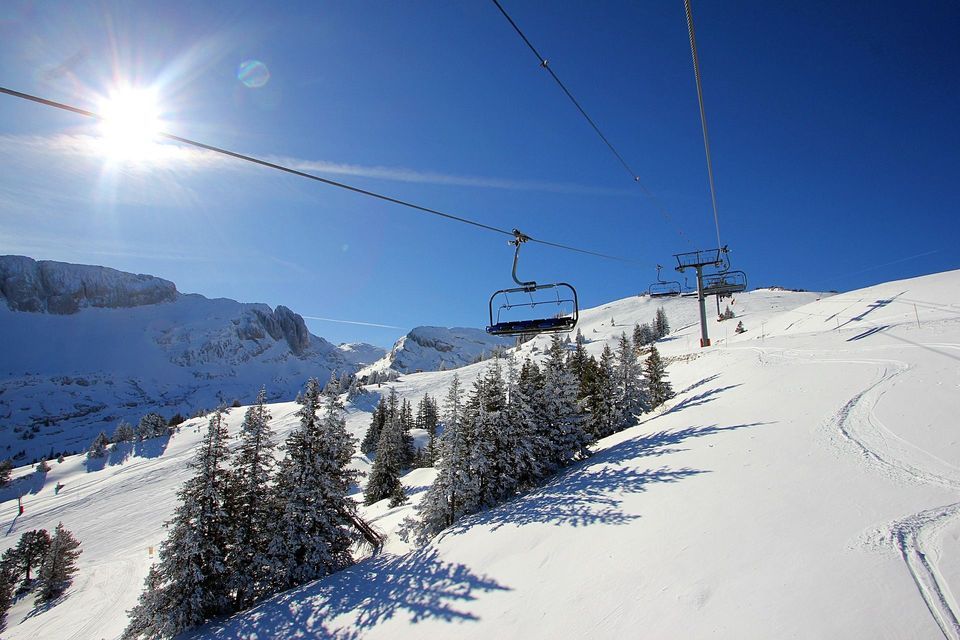 Un telesilla vacío asciende una ladera nevada con pinos abajo, bajo un sol brillante y un cielo azul claro.