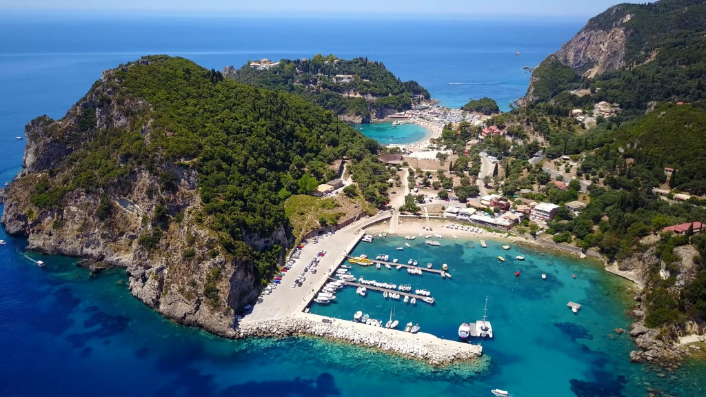 Una vista aerea di un porto con barche ormeggiate in acqua turchese, incastonato contro una costa verde e collinare.