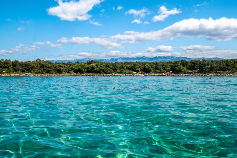 Acqua turchese e cristallina brilla in primo piano, con una spiaggia alberata e montagne in lontananza sotto un cielo blu con nuvole.