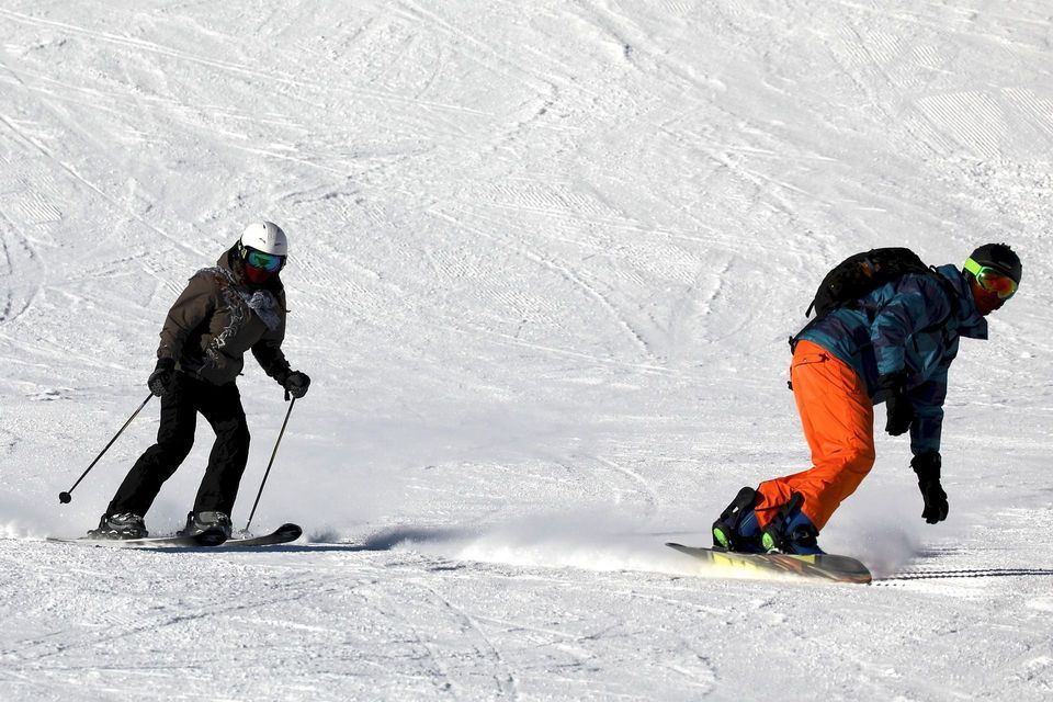 Un esquiador con casco blanco y un snowboarder con pantalones naranjas descienden por una amplia pista cubierta de nieve.