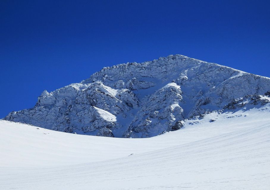 Un pico de montaña rocoso cubierto de nieve fresca bajo un cielo azul profundo y sin nubes, con una vasta extensión nevada en primer plano.