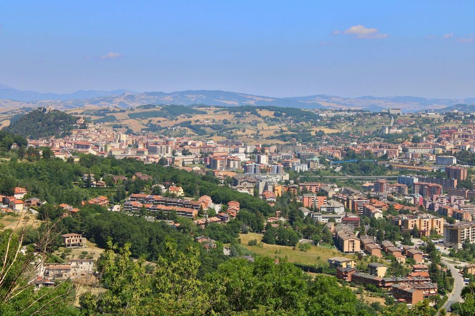 Una vista panoramica di una città estesa con edifici dai tetti rossi adagiati tra rigogliose colline verdi sotto un cielo azzurro brillante.