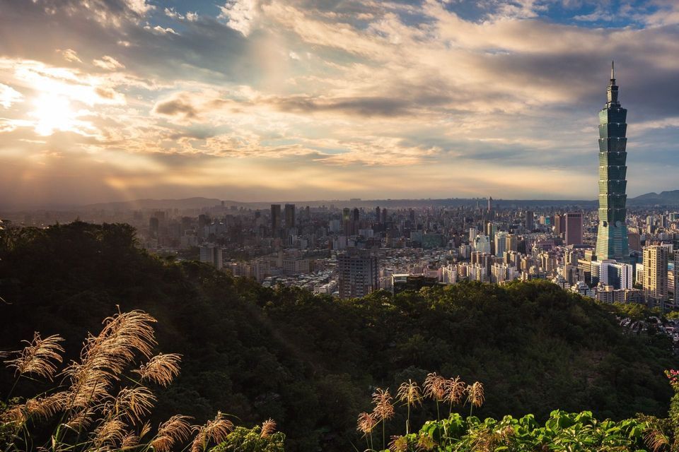 Eine weitläufige Stadtlandschaft mit einem hohen Wolkenkratzer, von einem üppig grünen Hügel bei Sonnenuntergang aus gesehen, mit goldenem Licht, das durch Wolken filtert.