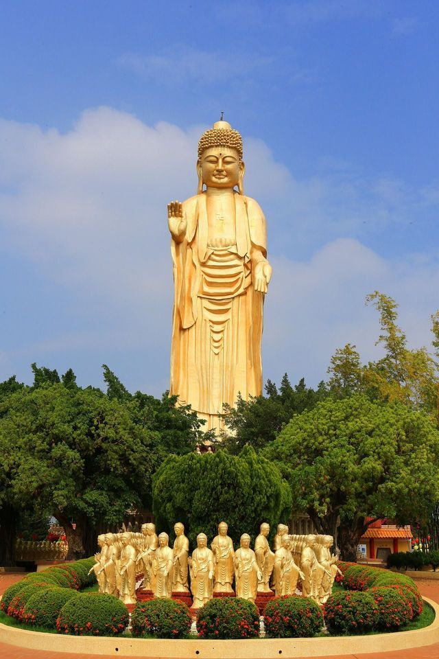 Eine riesige goldene Buddha-Statue ragt über einem üppigen grünen Garten mit vielen kleineren Statuen unter blauem Himmel.