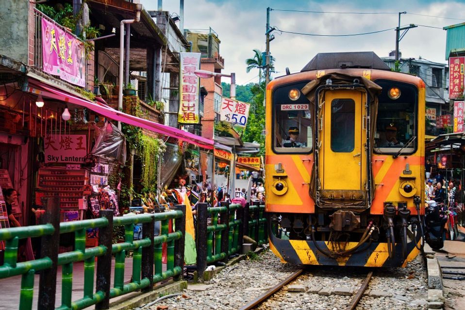 Ein gelb-orangefarbener Zug auf einer Eisenbahnstrecke, die durch eine belebte Marktstraße mit Geschäften und Menschen führt.