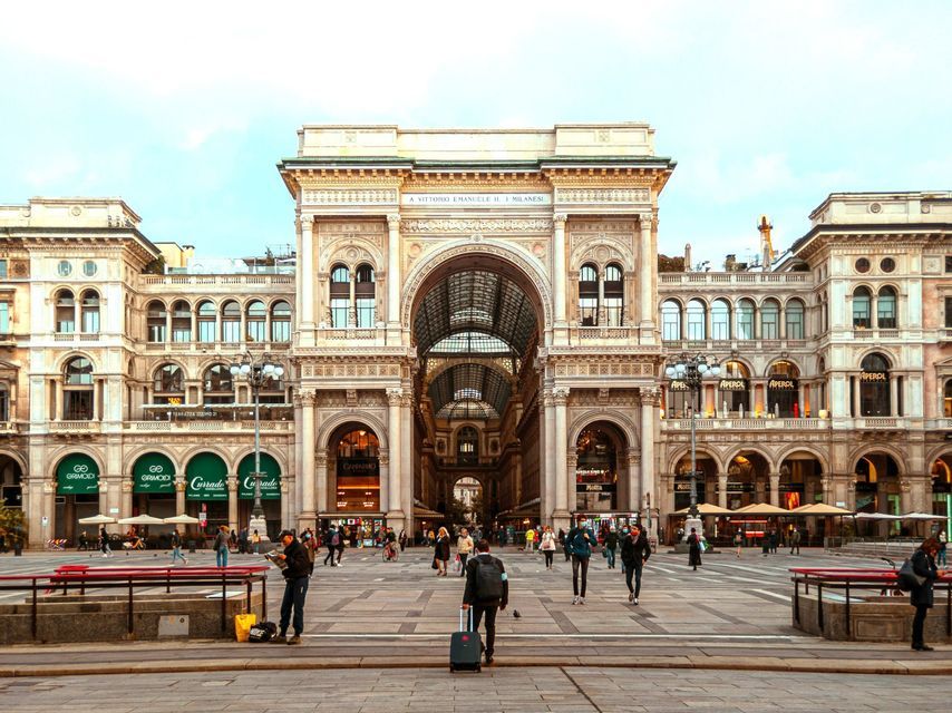 People walk across a large, paved square in front of the ornate facade and grand archway of a historic building.
