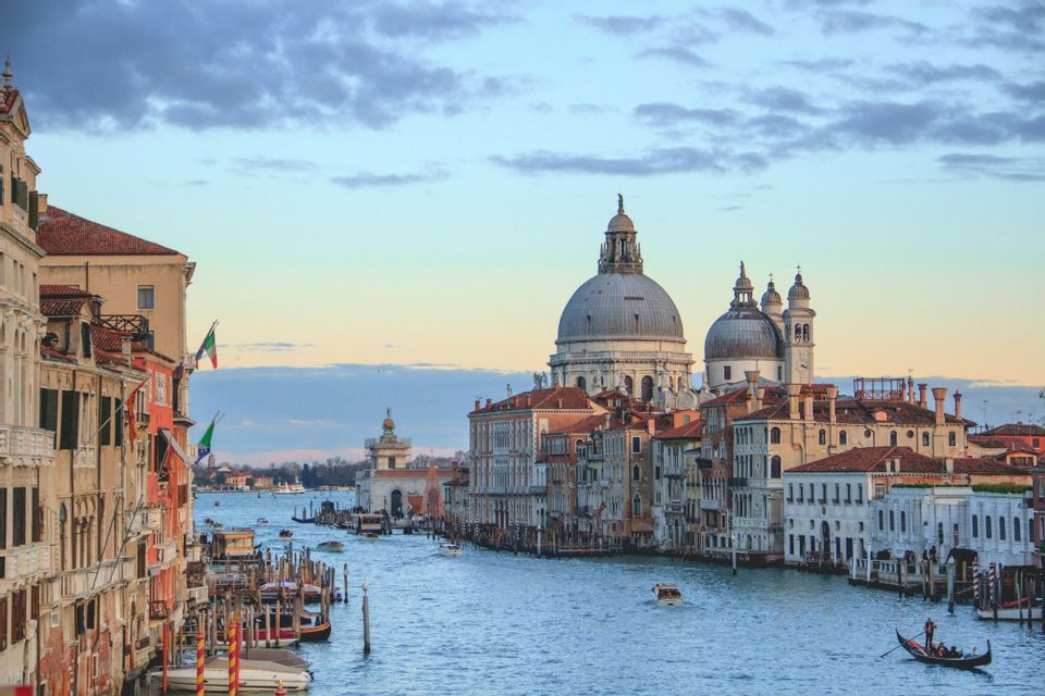 Boats and a gondola travel along a wide canal flanked by historic buildings, with a large domed basilica in the distance under a cloudy sky.