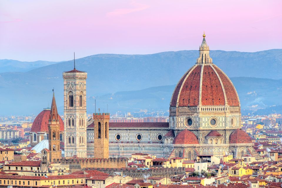 A large cathedral with a red-tiled dome and a tall bell tower overlooking a cityscape at dusk, with mountains in the distance.