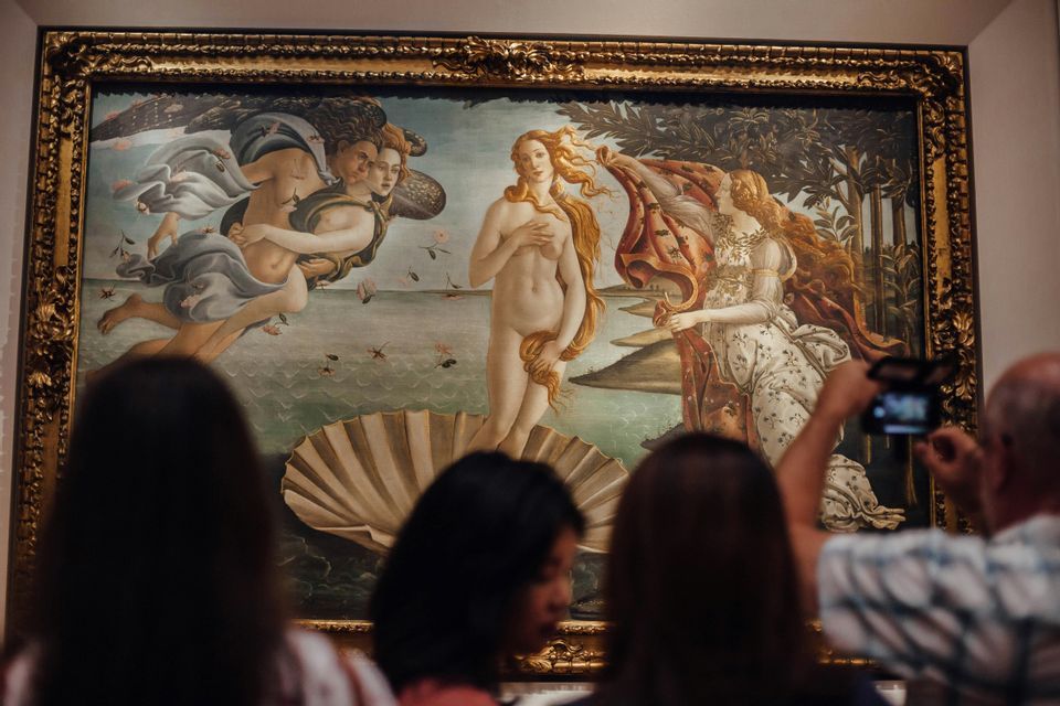 Museum visitors observe Botticelli's 'The Birth of Venus' in a large, gilded frame, with one person taking a photo.