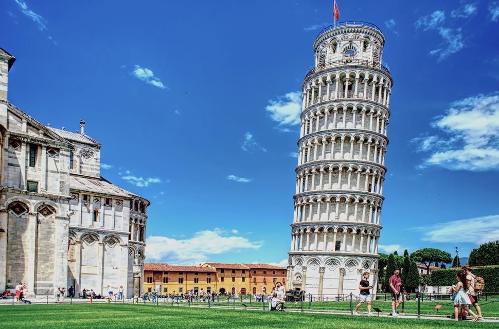 The Leaning Tower of Pisa and a cathedral stand on a green lawn with tourists walking by under a bright blue sky.