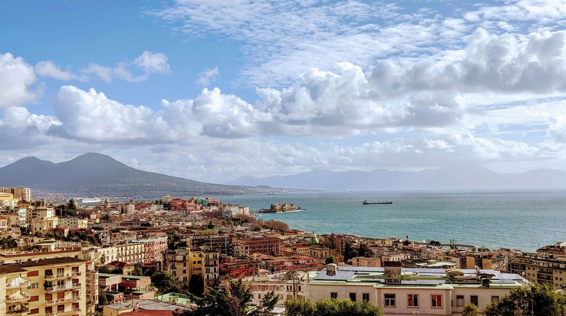 A panoramic view of a colorful coastal city, a bay, and a large volcano under a partly cloudy sky.
