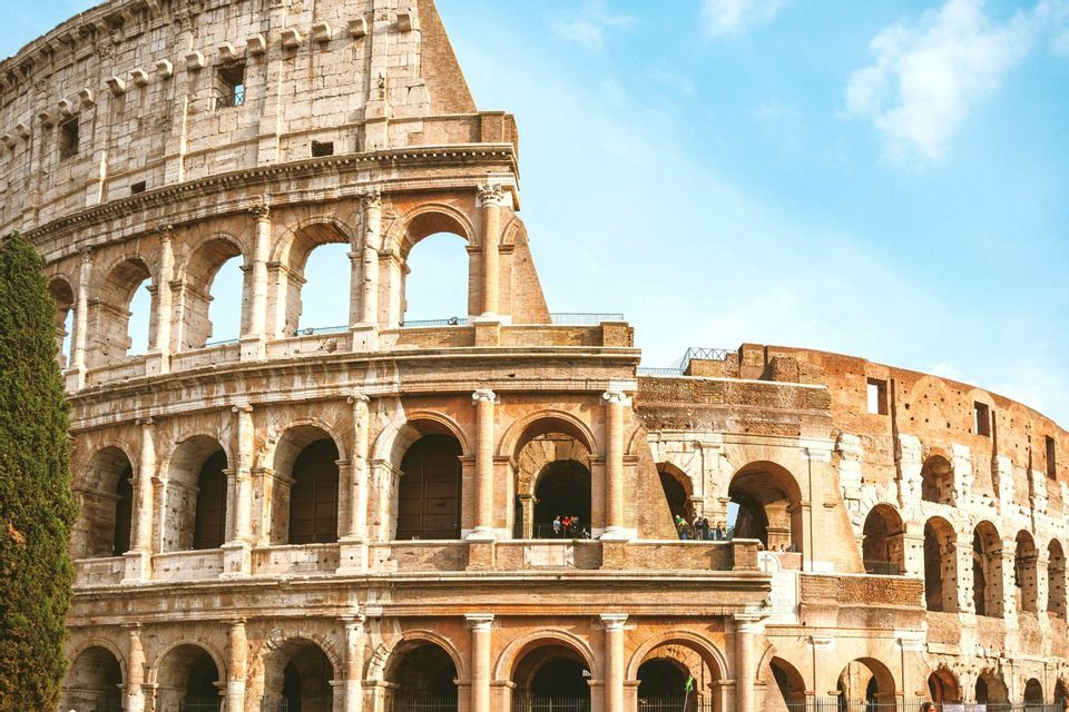 A sunlit, partial view of the ancient Roman Colosseum's tiered stone arches against a bright blue sky with light clouds.