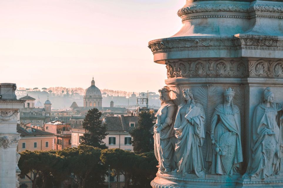 A close-up of carved marble statues on a monument, with a hazy city skyline featuring a large dome in the background.
