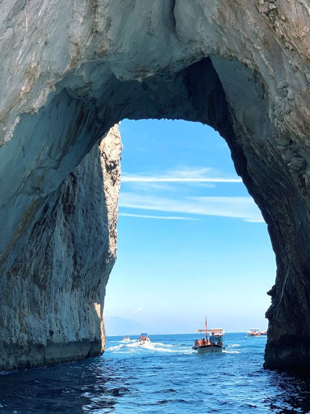 A WeRoad group trip on a boat sailing through a large rock arch on the open sea under a blue sky.