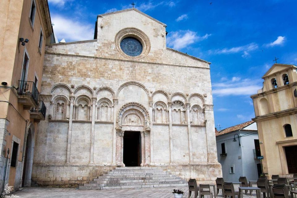 La facciata in pietra di una chiesa romanica con portali ad arco e un grande rosone, situata in una piazza cittadina.