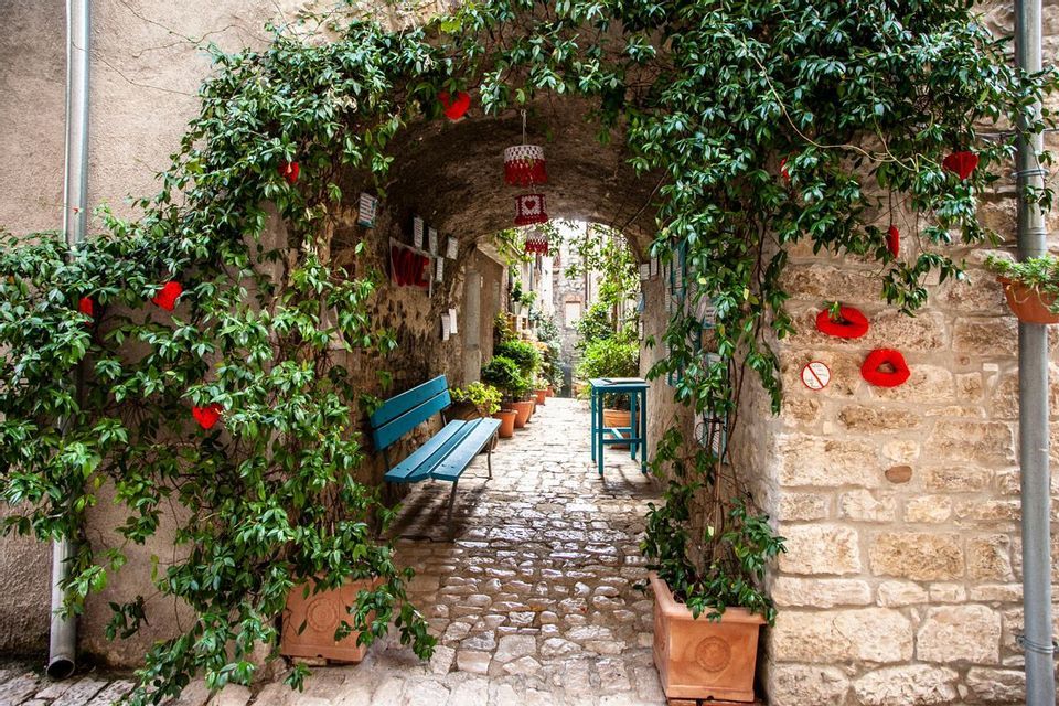 Una vista attraverso un arco in pietra coperto di viti in un vicolo stretto di ciottoli con una panchina blu e piante in vaso.