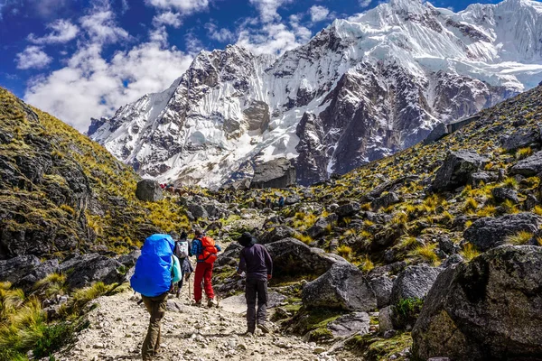 Un viaje en grupo de WeRoad camina por un sendero rocoso de montaña hacia un pico grande y cubierto de nieve bajo un cielo azul.
