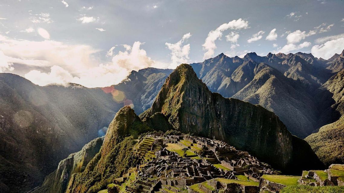Antiguas ruinas de piedra se asientan en una ladera montañosa verde y aterrazada, dominadas por picos escarpados bajo un cielo nublado y soleado.