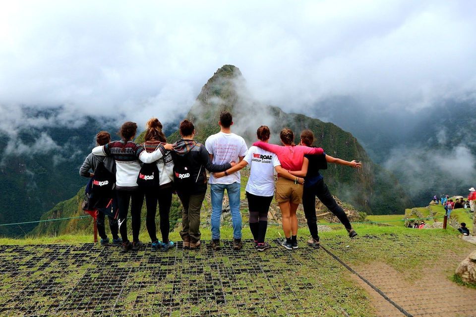 Un grupo de WeRoad, de pie y abrazados, en un mirador de hierba, contempla un pico de montaña cubierto de nubes.