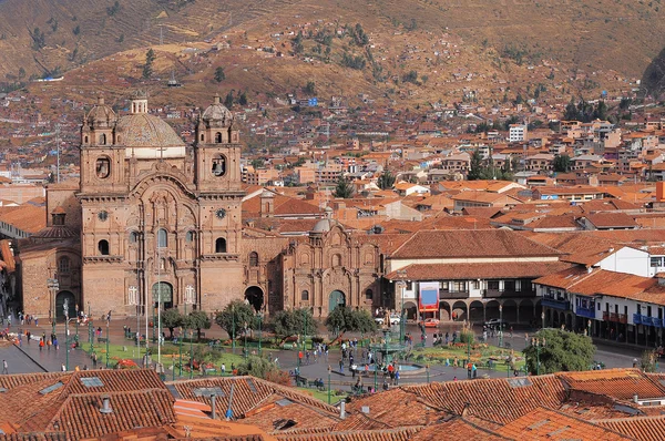 Una vista elevada de una histórica catedral de piedra en una concurrida plaza urbana, rodeada de edificios con tejados de teja roja en una ladera.
