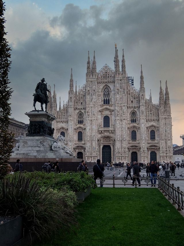 Una ornamentada catedral gótica vista desde una plaza con una estatua ecuestre y gente paseando bajo un cielo nublado.