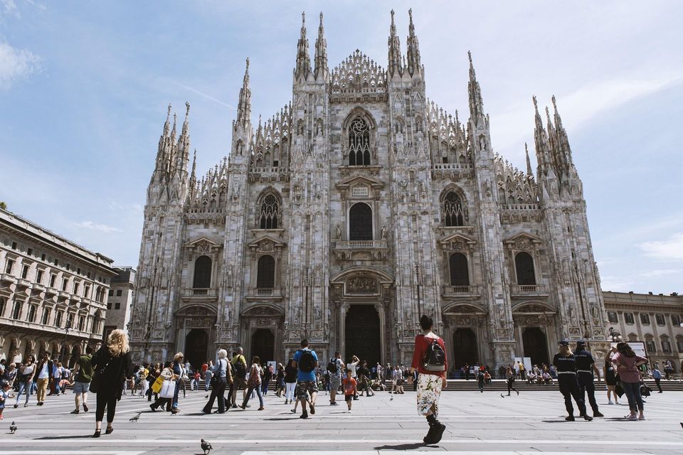 La ornamentada fachada de mármol blanco de una gran catedral gótica vista desde una animada plaza pública llena de gente en un día soleado.