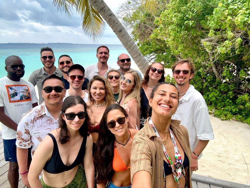 Un voyage de groupe WeRoad sourit pour un selfie sur une plage tropicale avec de l'eau turquoise et des palmiers en arrière-plan.