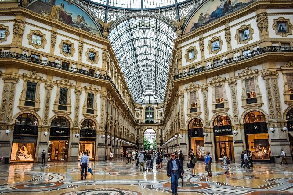 La gente pasea por una galería comercial ornamentada con un techo abovedado de cristal, intrincados suelos de mosaico y escaparates de lujo.