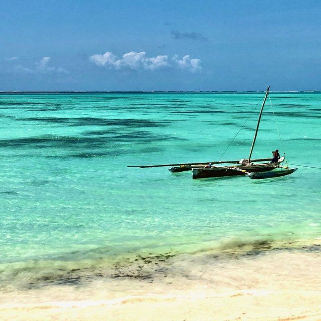 Una persona sentada en una canoa tradicional de balancín flota en aguas claras y turquesas cerca de una playa de arena bajo un cielo azul.