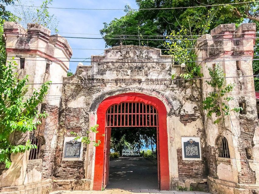 Un portone di fortezza in pietra logorata, con un ingresso ad arco rosso vivo e aperto, invaso da piante verdi.