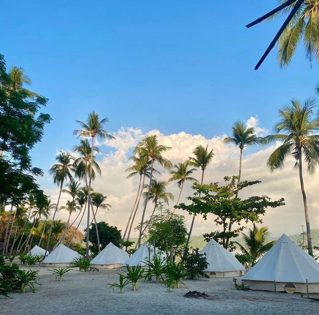 Tende glamping bianche su una spiaggia sabbiosa, circondate da alte palme sotto un cielo parzialmente nuvoloso.