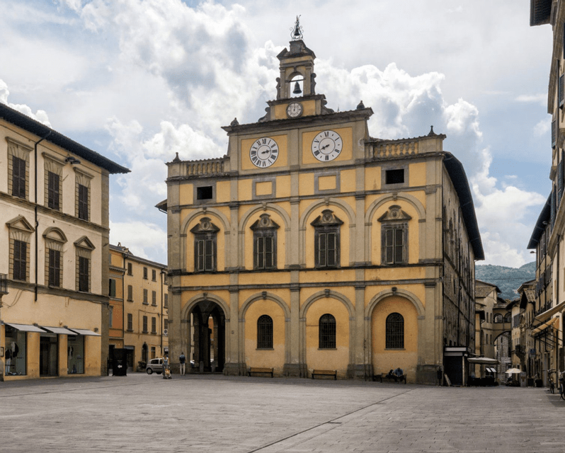 Un edificio comunale giallo e ornato con una torre dell'orologio si erge in una grande piazza cittadina pavimentata sotto un cielo nuvoloso.