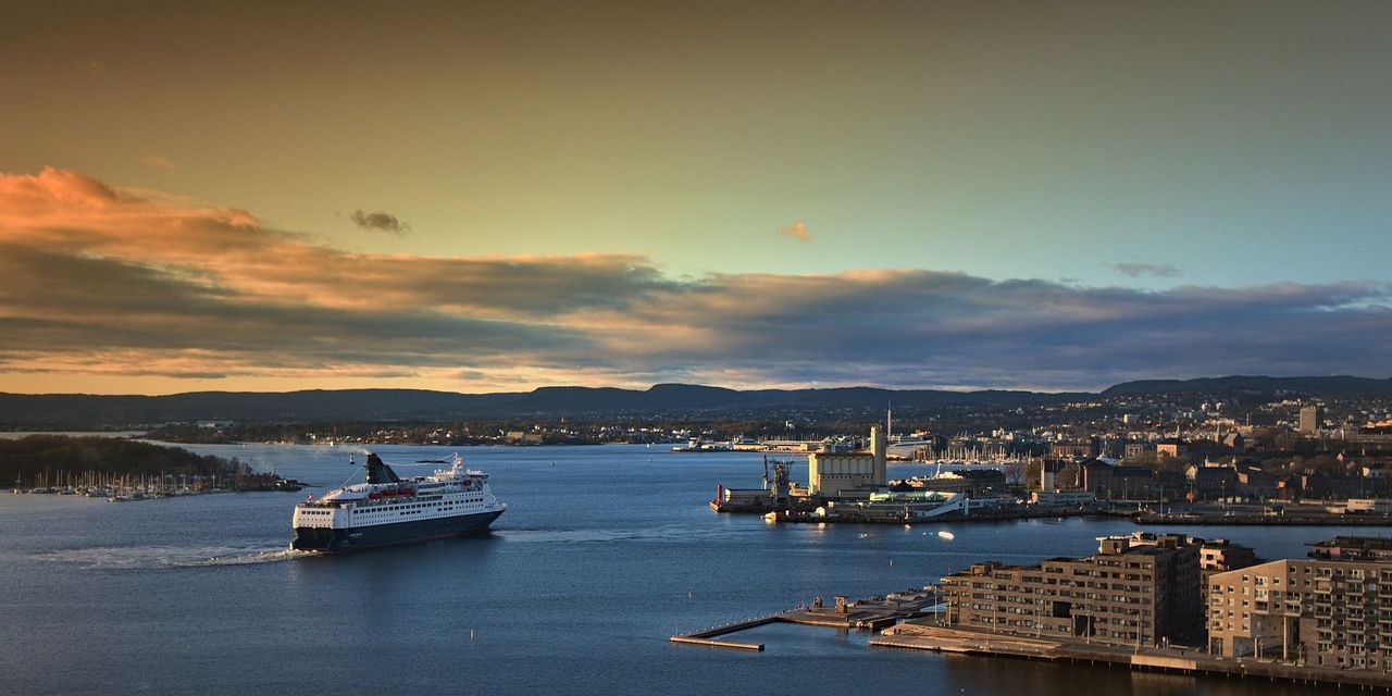 A large ferry sails across a bay towards a harbor city at sunset, with distant hills under a cloudy sky.