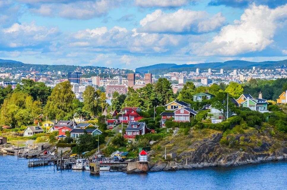Colorful houses on a rocky, tree-covered coastline with a city in the background under a blue sky with white clouds.