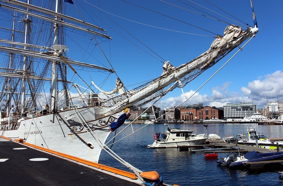 The bow of a large white tall ship with intricate rigging docked in a city harbor under a bright blue sky.