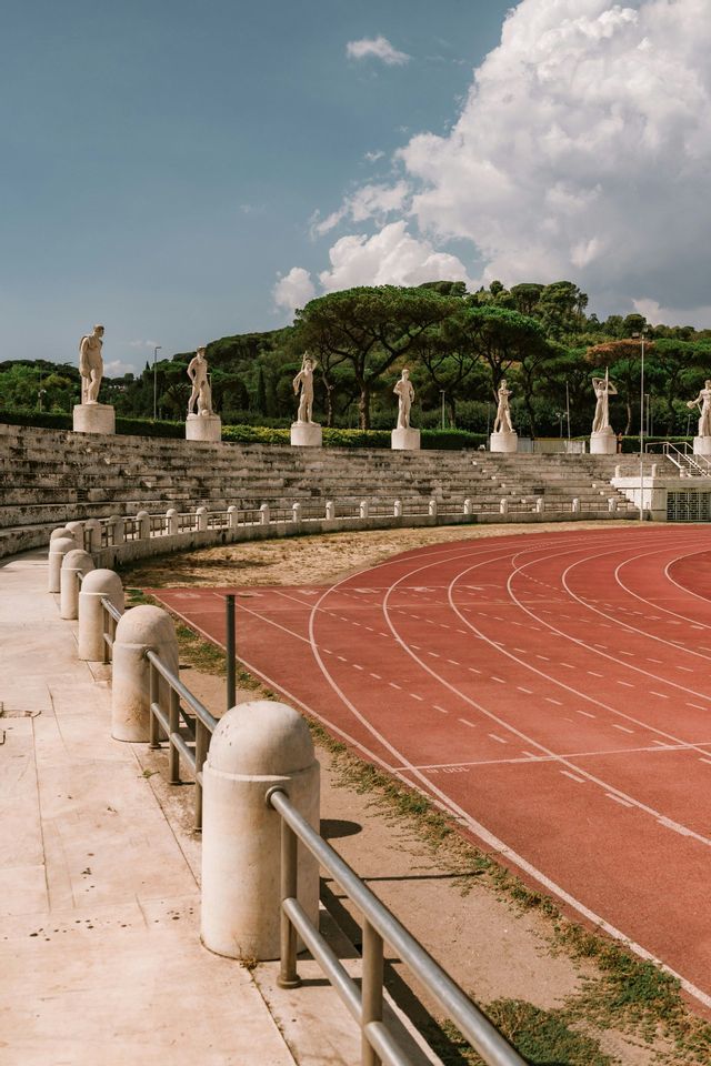 Una fila di statue classiche si erge su gradinate di pietra che si affacciano su una pista di atletica rossa in uno stadio all'aperto.