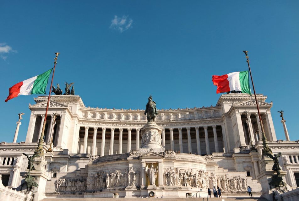 Due bandiere italiane sventolano sopra un grande monumento in marmo bianco con colonne e una statua equestre sotto un cielo azzurro chiaro.