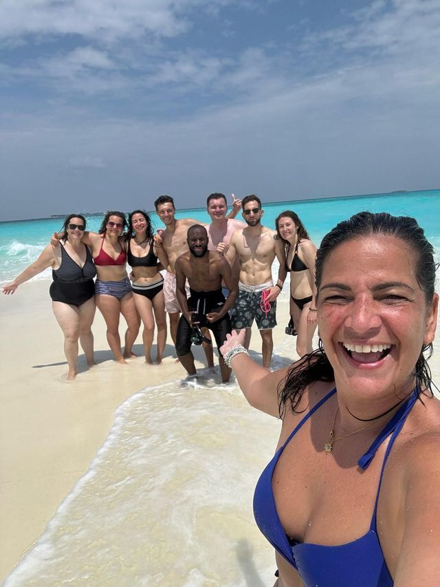 Une femme en bikini bleu prend un selfie souriant avec son voyage de groupe WeRoad sur une plage de sable au bord de l'océan turquoise.