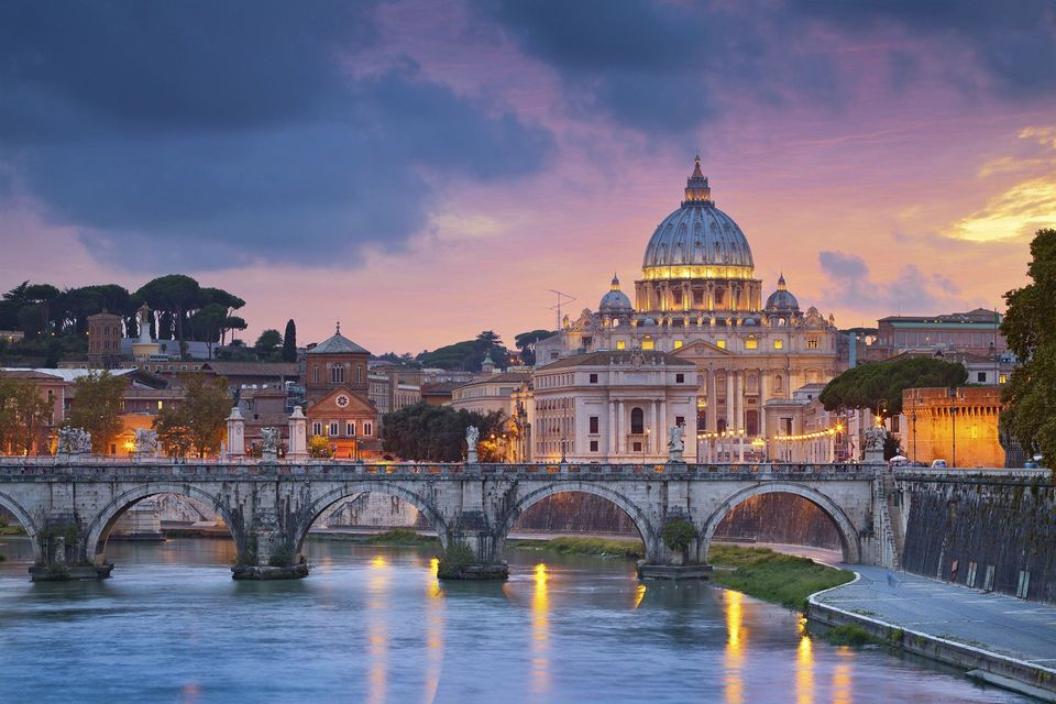 Una basilica a cupola illuminata e un ponte ad arco in pietra che attraversa un ampio fiume al crepuscolo, sotto un cielo nuvoloso e colorato.