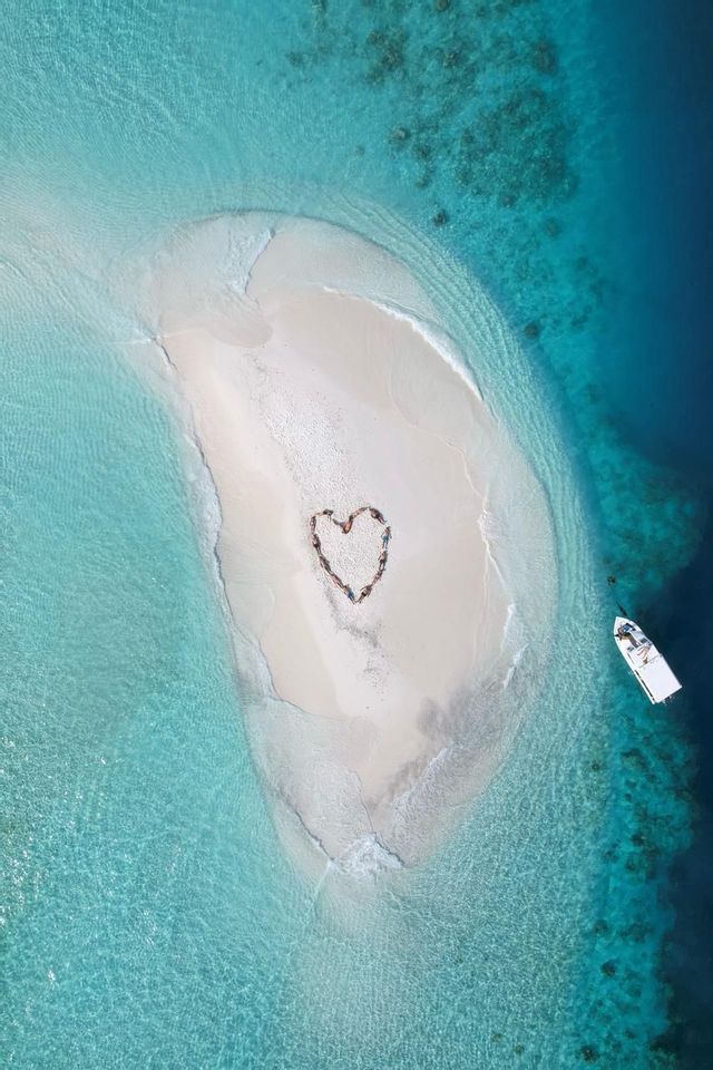 Vue aérienne d'un groupe WeRoad en voyage, allongé sur une île de sable blanc et formant un cœur dans l'océan turquoise.