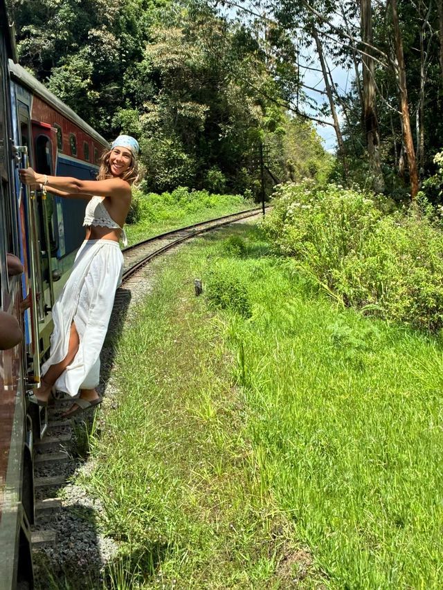 Une femme souriante vêtue de blanc et portant un bandana se penche hors d'un train en mouvement alors qu'il traverse une forêt luxuriante et verte.