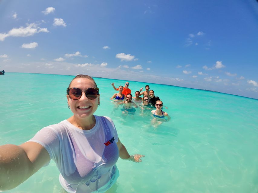 Une femme prend un selfie avec son groupe de voyage WeRoad, toutes souriantes dans l'eau claire et turquoise de l'océan sous un ciel bleu.