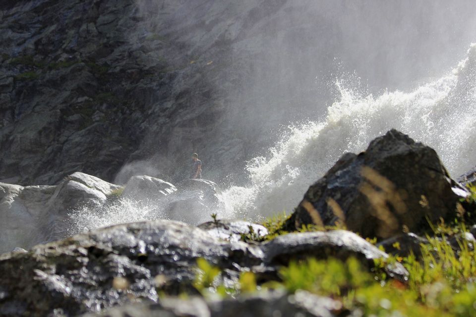 Eine Person steht auf einem großen Felsen neben einem mächtigen, spritzenden Wasserfall, der eine Nebelwolke erzeugt.