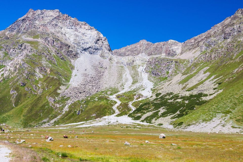 Ein weites, grünes Alpental am Fuße hoher, felsiger Berge unter einem klaren, tiefblauen Himmel tagsüber.