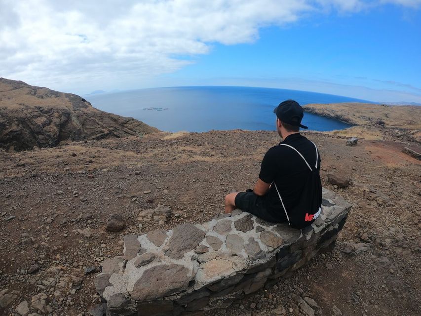 Un uomo visto di spalle siede su un basso muretto di pietra su una scogliera, osservando l'oceano in una giornata parzialmente nuvolosa.