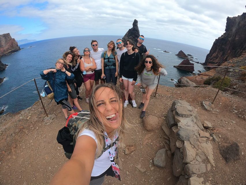 A WeRoad group trip takes a wide-angle selfie on a cliffside overlooking the ocean and rocky formations.